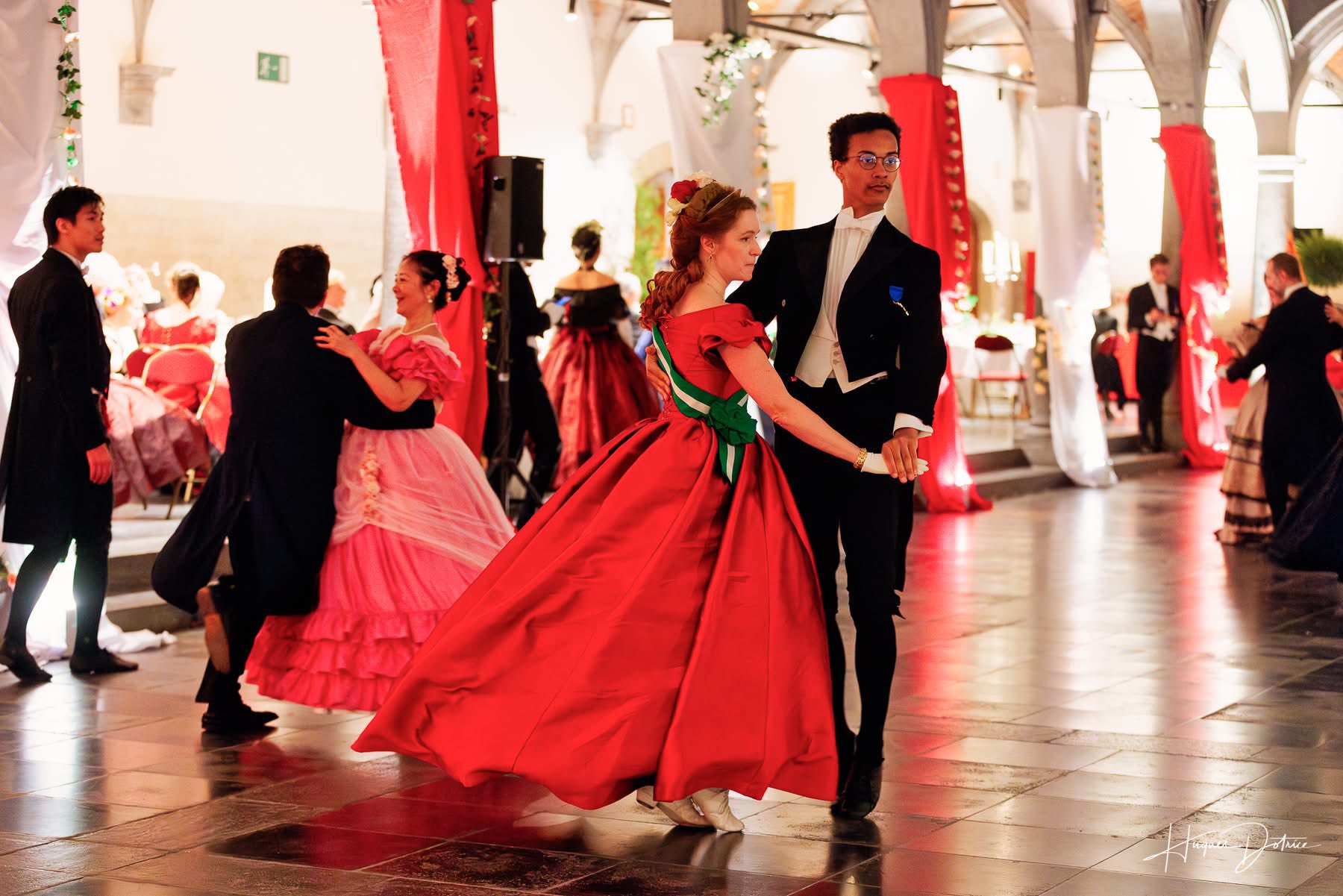 Un couple en costumes historiques au bal, avec une danseuse en robe rouge au premier plan
