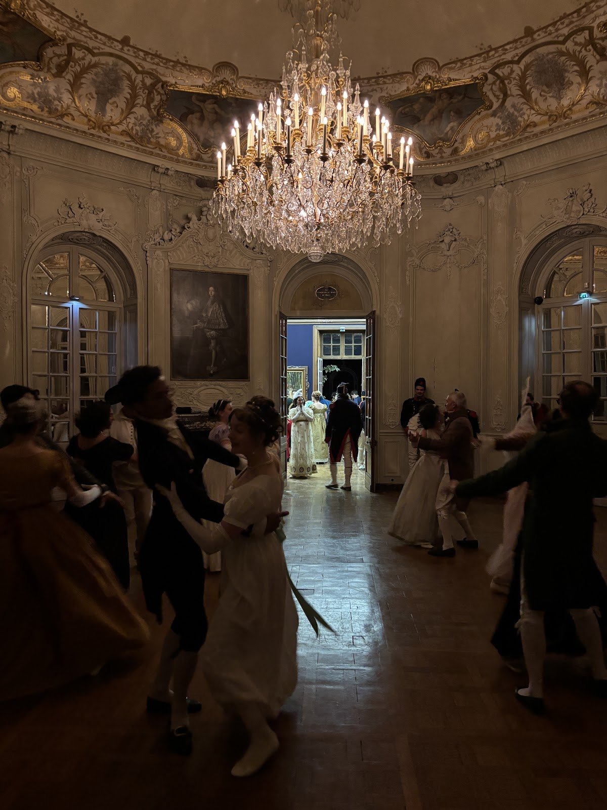 Couples en costumes Premier Empire dansant sous un grand lustre dans le salon d’une dépendance du Château de Chantilly