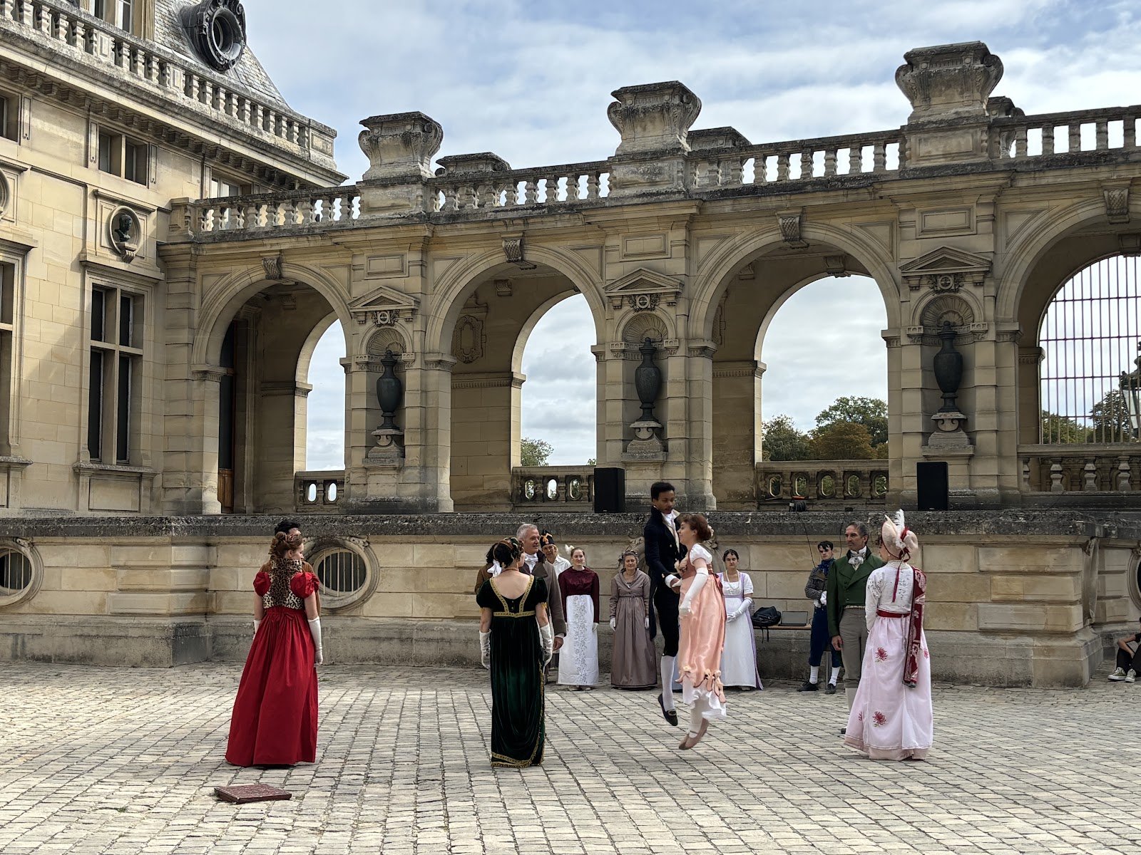 Danseuses et danseurs en costumes Premier Empire se tenant par la main devant les arcades de la cour de Chantilly