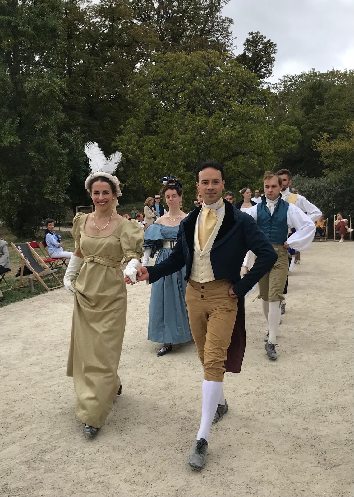 Un couple en costumes historiques au premier plan, suivi d’autres danseurs dans le parc de la Vallée aux Loups